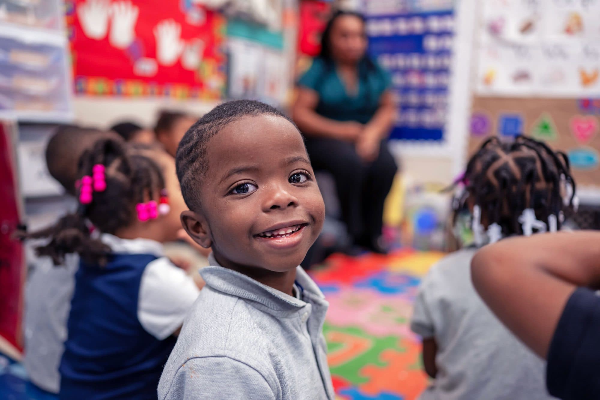 Smiling young student sitting on a colorful classroom mat with classmates.